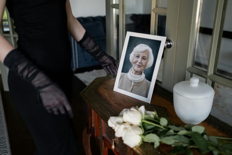 operatrice funebre accanto a una bara, con sopra la foto di una signora, un’urna cineraria bianca e rose