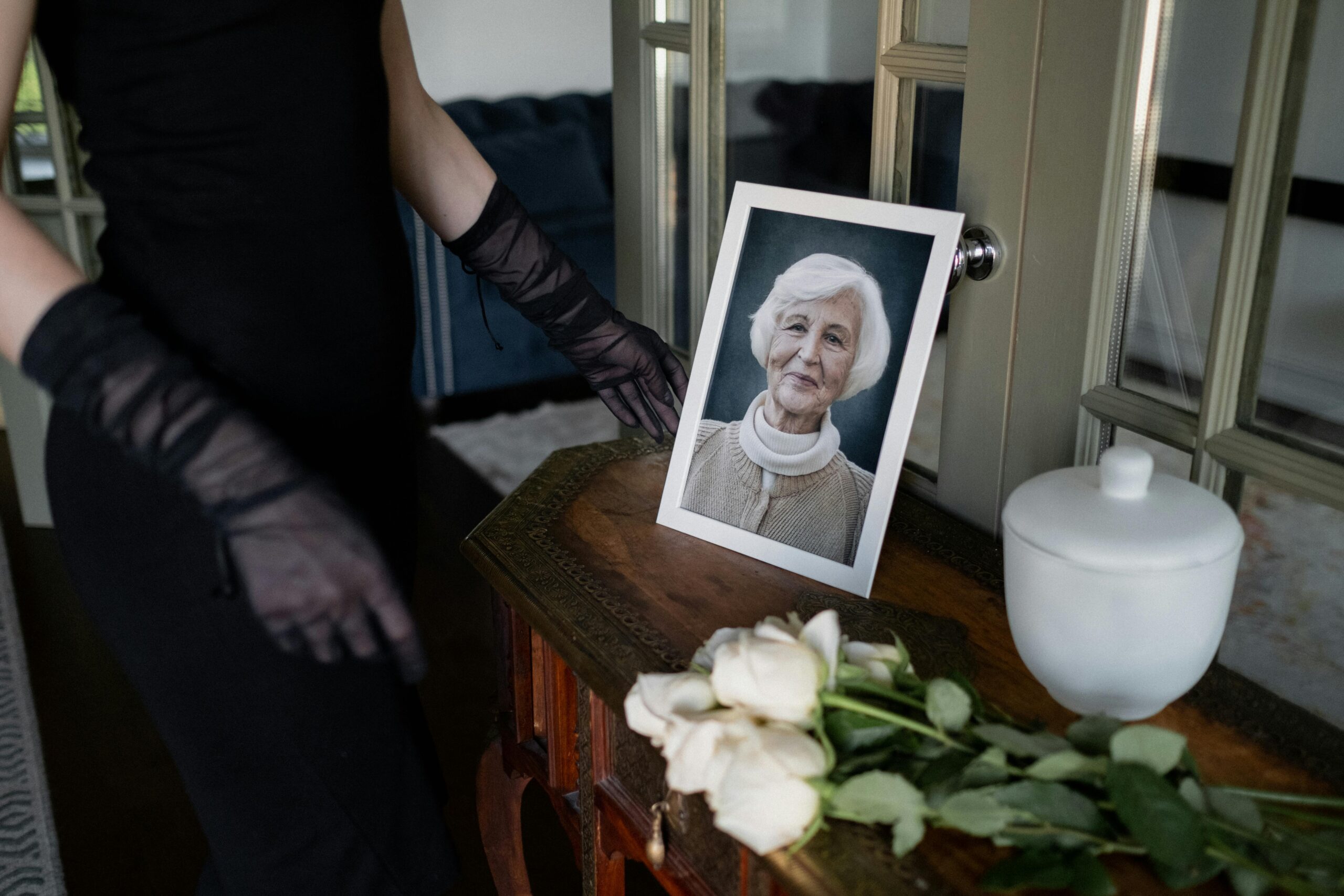 operatrice funebre accanto a una bara, con sopra la foto di una signora, un’urna cineraria bianca e rose